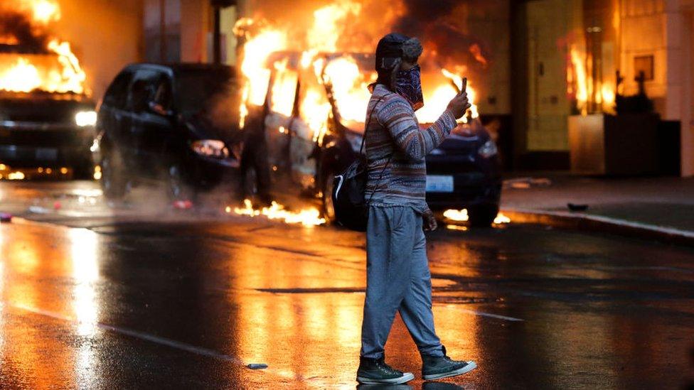 Man uses his phone to record events ahead of him. Burning cars in the background.