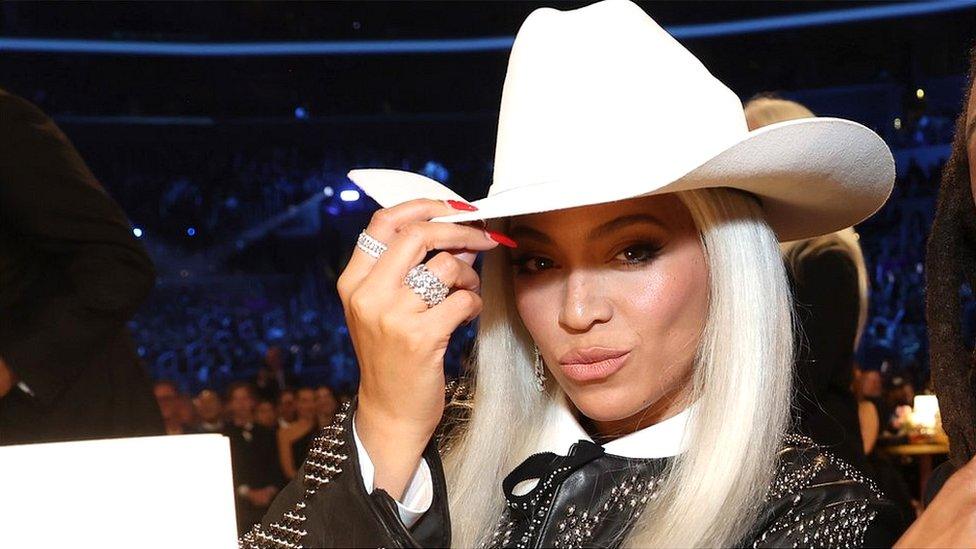 Beyoncé pictured at the 2024 Grammys wearing a white cowboy hat, pouts at the camera. The singer has bleached blonde hair worn loose and doffs her cap with her right hand, her nails painted bright red and chunky silver rings on her fingers. She wears a studded leather jacket and matching studded bow tie over a white shirt.