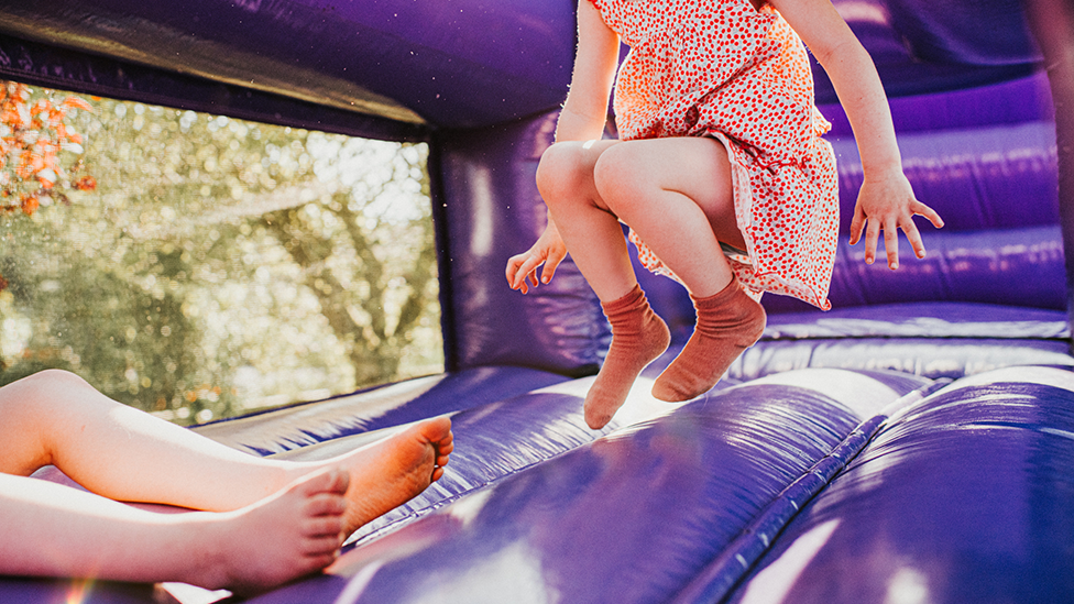 The legs and torso of a girl jumping in mid air while wearing orange socks and a dotted dress on a purple inflatable castle. The legs of another child can bee seen in the bottom right-hand corner