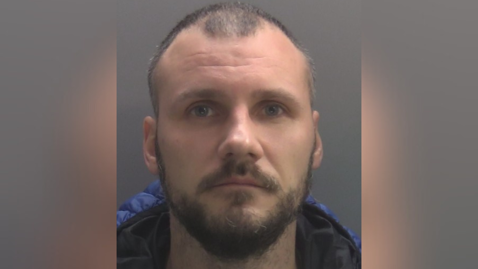A mugshot of Donatas Venclovas. A white man in his 30s, with a short, dark, untidy beard and moustache and a receding hairline.
He is wearing a blue, hooded, padded coat. He appears expressionless.