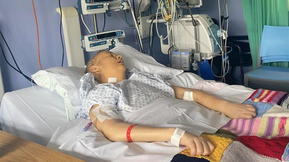 Claire Boyd, who is unconscious, shown in a hospital bed. She is attached to lots of different machines.