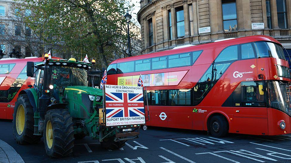 A green tractor with a protest banner and Union flags on the front next to a red  bus on Whitehall in London.