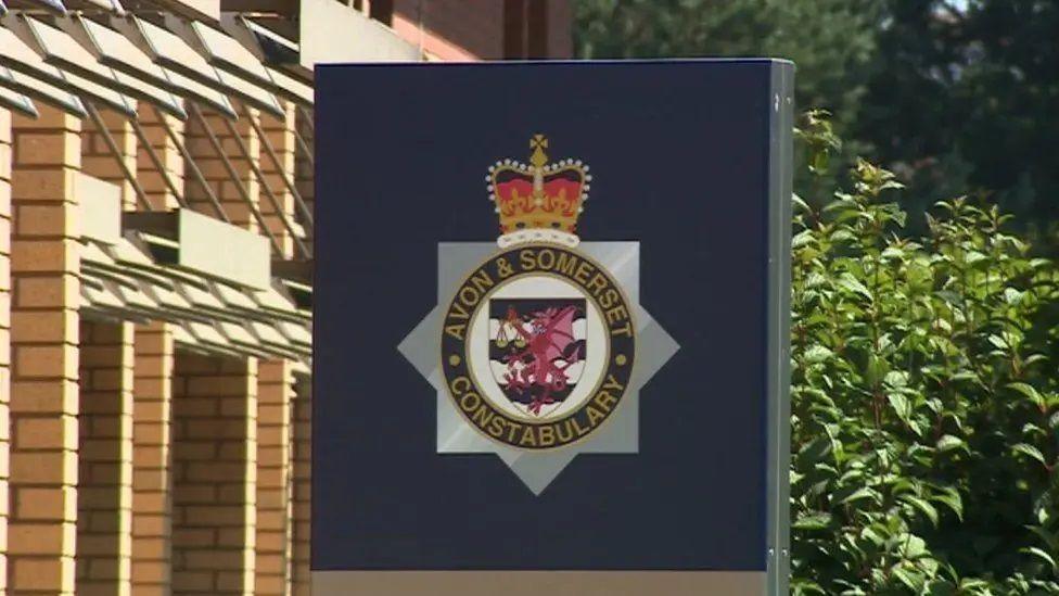 The outside of Avon and Somerset Police's headquarters. The picture is a close up of the sign outside which shows the force logo. A light-brick building is seen in the background, and a large green bush. 