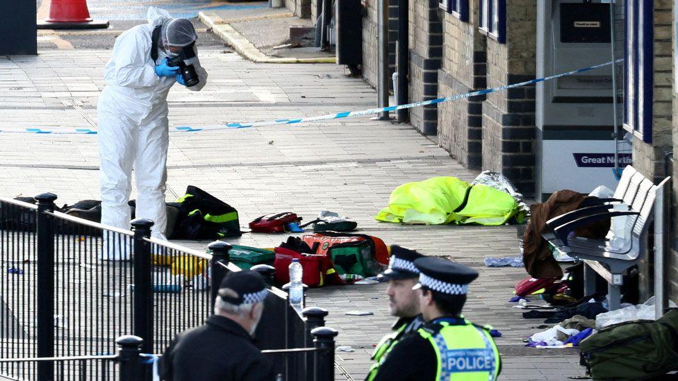 A forensic officer in a white suit bending over and pointing their camera at the platform. On the platform are scattered possessions. Three police officers are in the foreground.