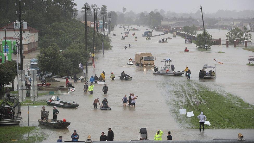 Image shows a flooded street in Houston with rescue boats and people wading through knee-deep water.