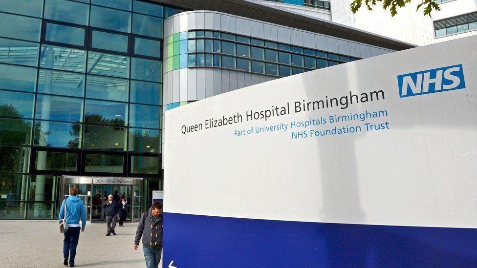 An outside view of Queen Elizabeth Hospital in Birmingham with windows in the background and the hospital visible inside with pillars and an entrance lobby.