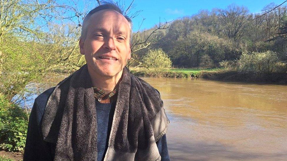 A man standing by a river with his towel round his neck