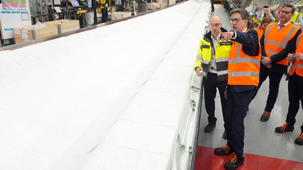 Ed Miliband at the Siemens Gamesa factory in high visibility jacket points as he stands next to a white turbine blade surround by people also in high visibility jackets