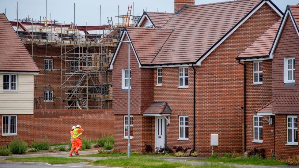 Two contruction workers in orange hi vis walk in front of a red brick house on a newly built estate