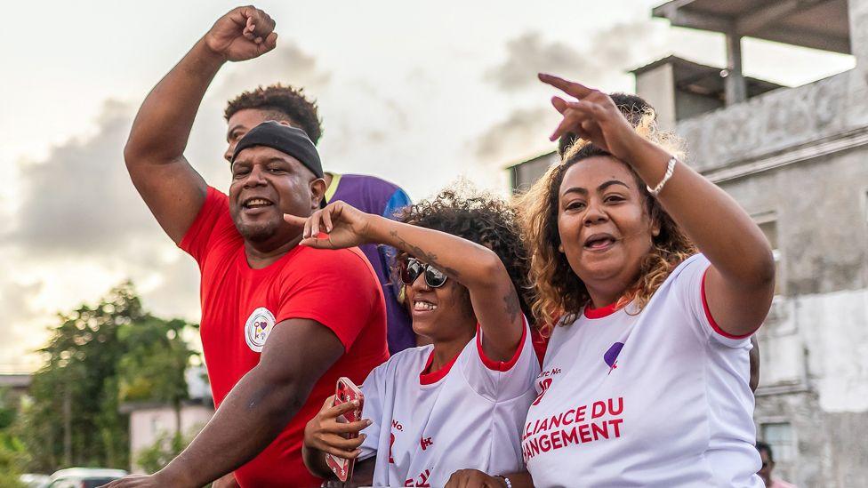 Three opposition supporters in red and white T-shirts wave their hands in the air in celebration.