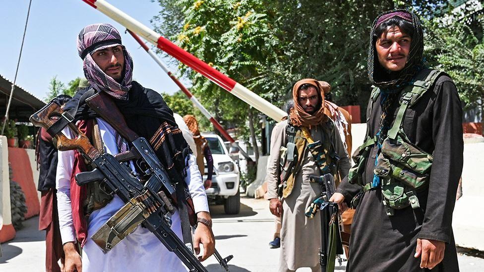 Three men wearing headdresses, standing near a rising gate for vehicles. They are all heavily armed, at least two of them with machine guns.