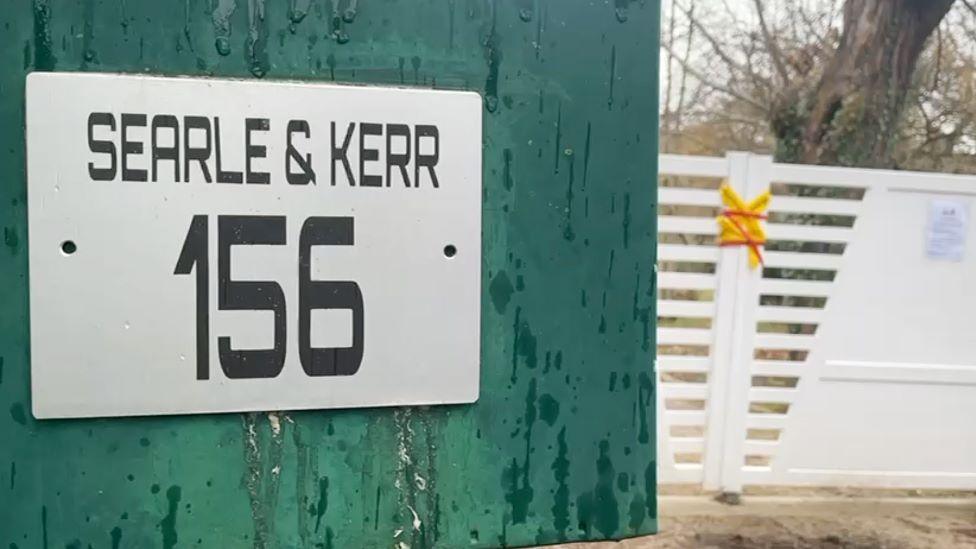 In the foreground there is a teal-coloured post-box that says "Searle & Kerr 156". Bright yellow and bright red police tape covers a white gate in the background.