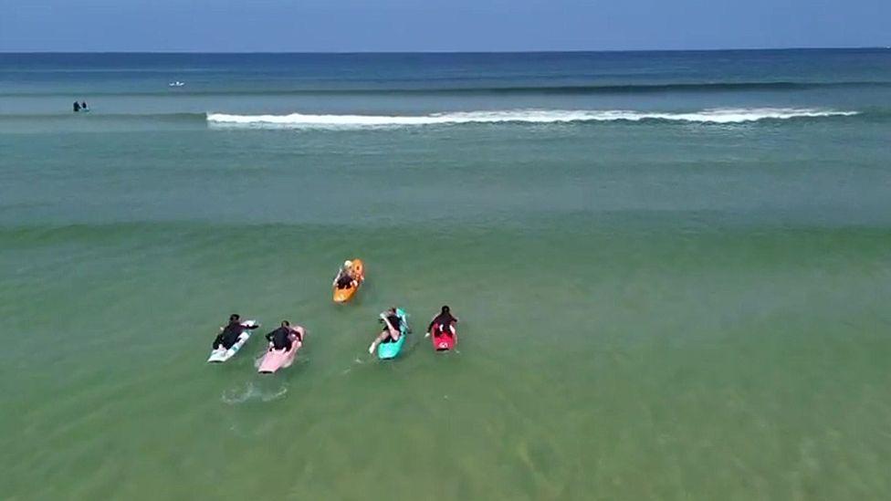 An aerial shot of four paddle boards in yellow, multi-coloured, pink, blue and red set off across the waves at Hayle.