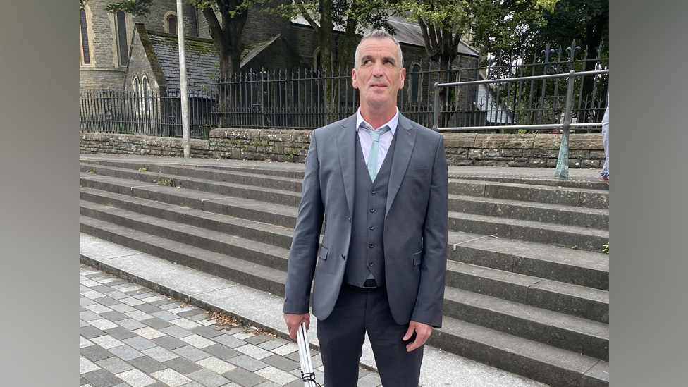 A blind man holding a folded up white cane with a black cord wrapped around it. He is wearing a grey two piece suite with a white collar shirt underneath a wait coast and a light sky blue slim tie. He is standing in front of a gothic looking church