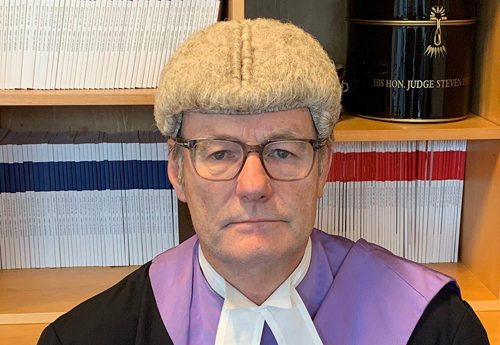 Judge Steven Parker, wearing wig and gown, looking at the camera in front of a bookshelf