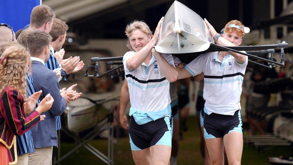 Rowing team with upturned boat on their shoulders - crowd clapping as they pass