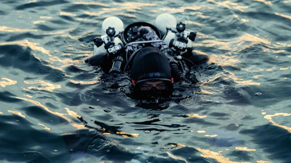 A diver in full underwater gear who is largely submerged beneath the water