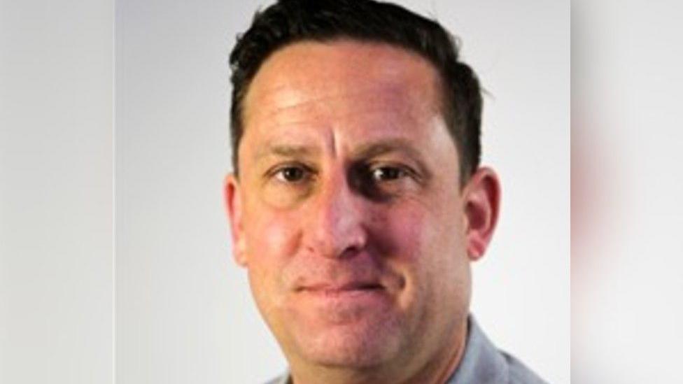 A headshot of a man wearing a red top and a grey jacket.