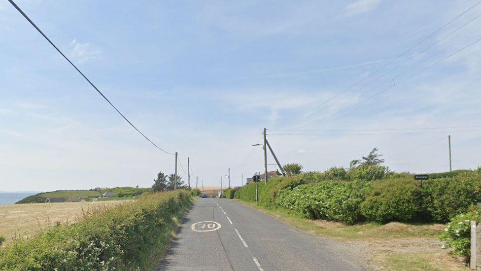 A rural road with trees and bushes down the side and a 30mph sign in the middle of the carriageway on a blue sunny day
