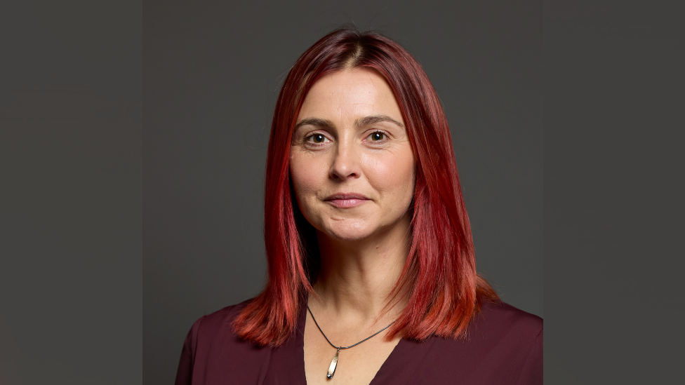 A head shot of Melanie Onn. She has shoulder-length red hair an dis wearing a black necklace with a silver charm. She is also wearing a burgundy blouse an dis looking at the camera. The background is grey. 