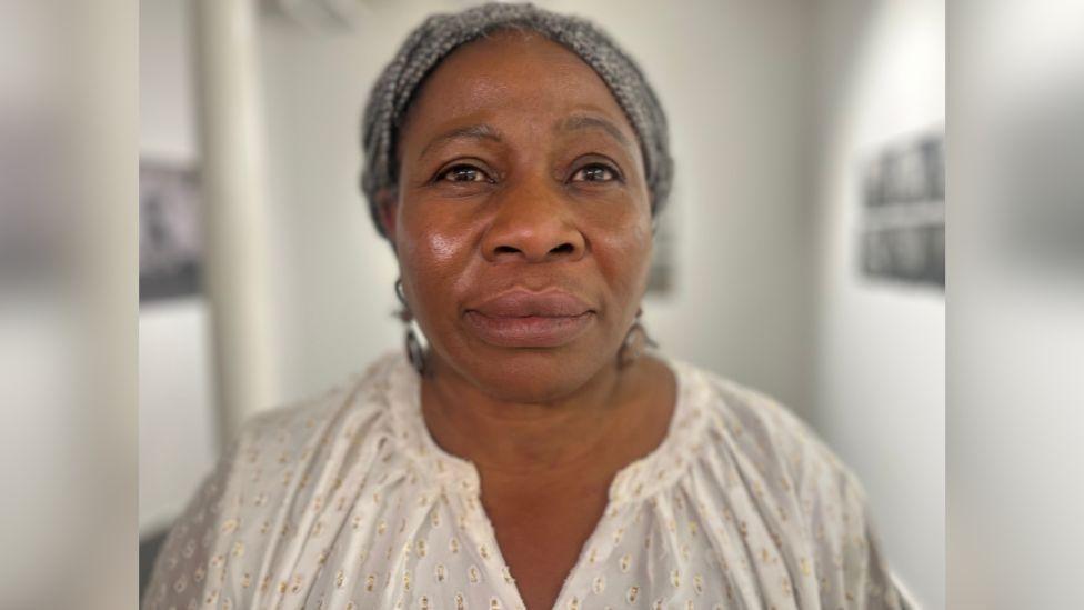 A woman stands and stares at the camera. She has braided grey hair and wears a white top.