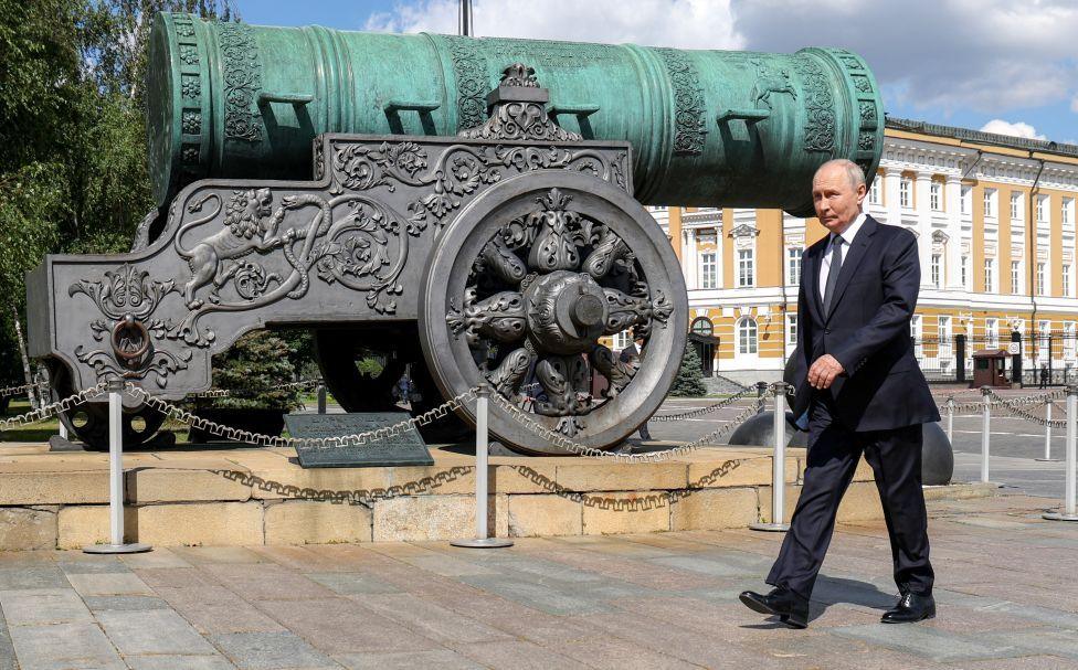 Putin walks past a Tsar Cannon monument in Moscow. He is wearing a dark suit, blue tie and white shirt. The cannon is very ornate and situated on a pedestal.