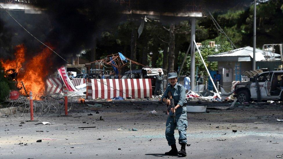 An Afghan policeman stands guard as smoke and flames rise from the site of a huge blast struck near the entrance of Kabul's international airport - 10 August 2015