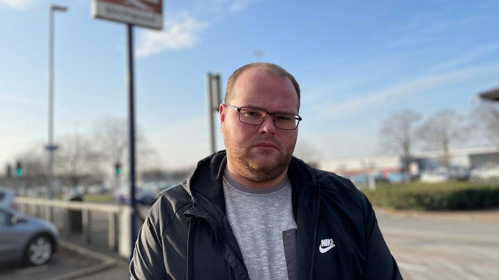 A man with glasses looks at the camera with a sombre expression on his face