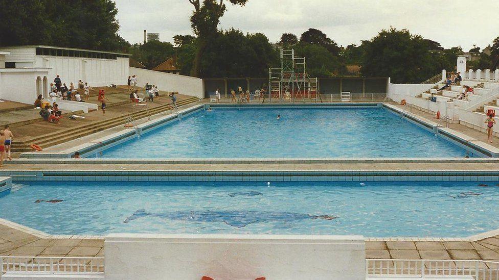Broomhill lido pictured in 1969. Blue tiles in the shape of a whale can be seen at the bottom of the pool. There are people sitting around the edge of the pool.