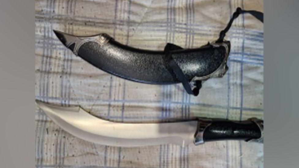 A machete knife with a curved blade and black handle, lies on a blue and white checked fabric background, next to its black sheath