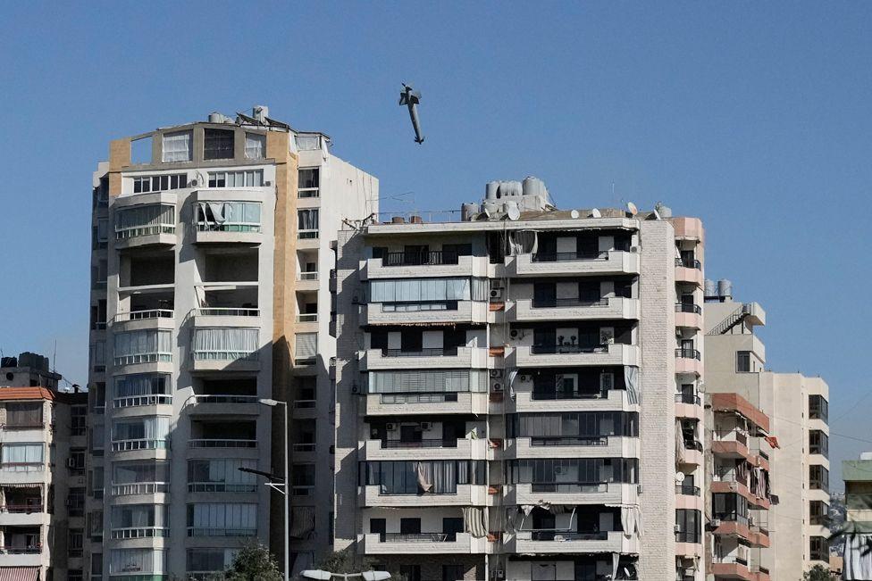 A missile flies towards a concrete grey building with balconies, the sky is clear and blue