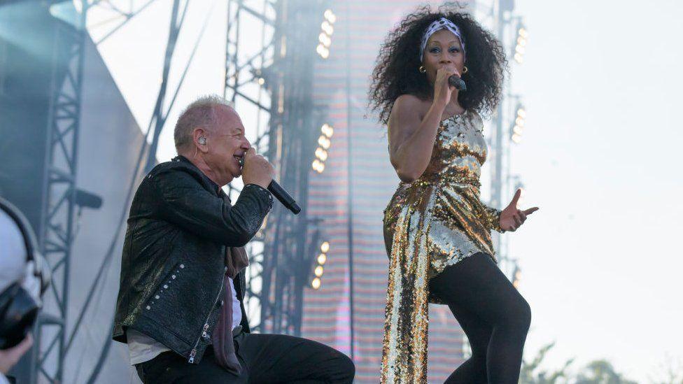 Jim Kerr of Simple Minds crouched down with a singer in a gold sparkly dress in front