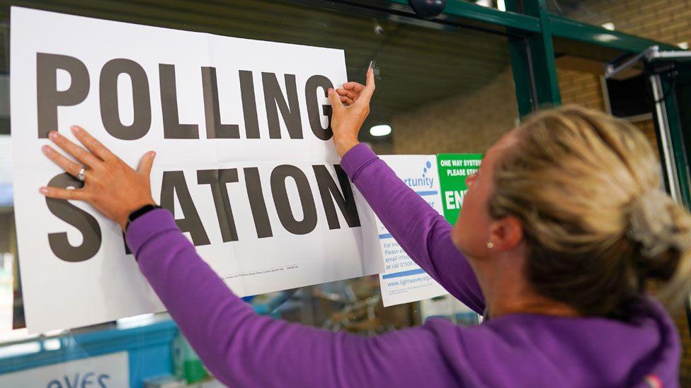 An official with blonde hair and a purple top attaches a sign to a polling station as voting begins in the by-election on 23 June 2022 in Wakefield, England.