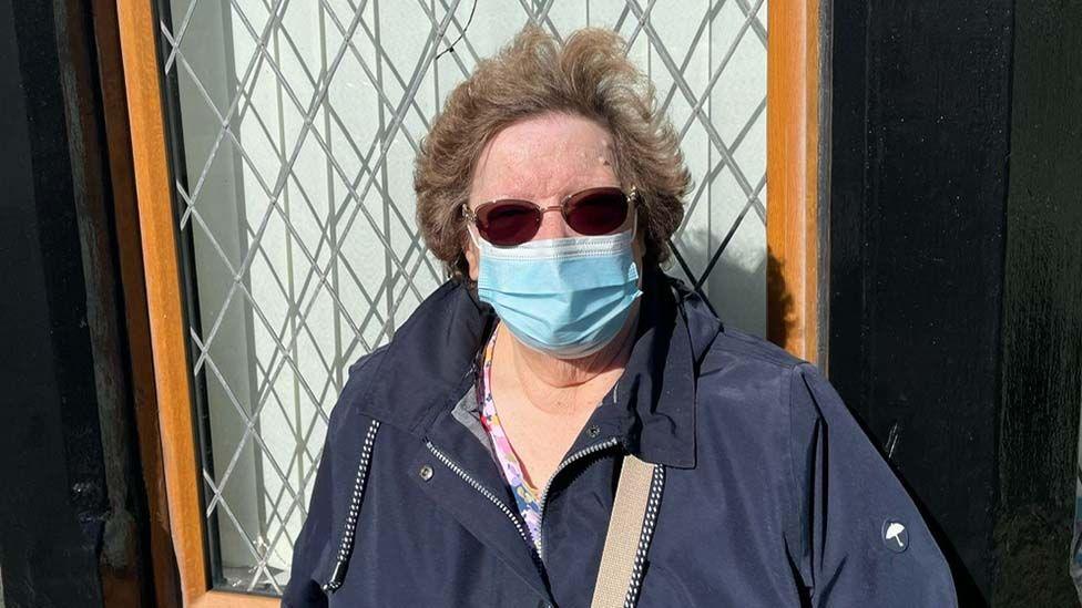 Elizabeth Lamb standing in front of a house window. She is wearing a navy blue coat and has short brown hair and sunglasses. She is wearing a blue face mask