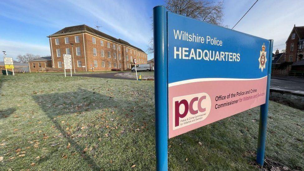 The Wiltshire Police Headquarters on a frosty morning. There is a large rectangular brick building in the background and a blue sign in the foreground. The sky is a pale blue with wispy clouds, and the trees are bare of leaves.