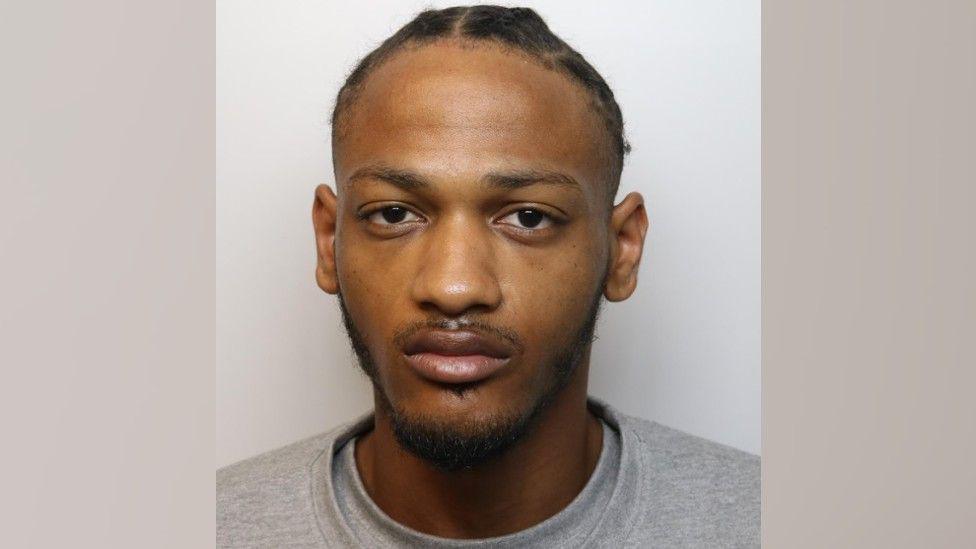 A custody picture of Maurice Jones. He is wearing an grey t-shire and jumper, and his hair is pulled back in braids. 