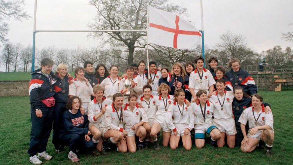 The England team pose for a victory photo on a green field in front of white rugby posts. The 15 players are wearing white shirts and shorts with half standing in the back row and the rest kneeling in the front row. Coaches and support staff, wearing blue tracksuits, are also in the picture.
