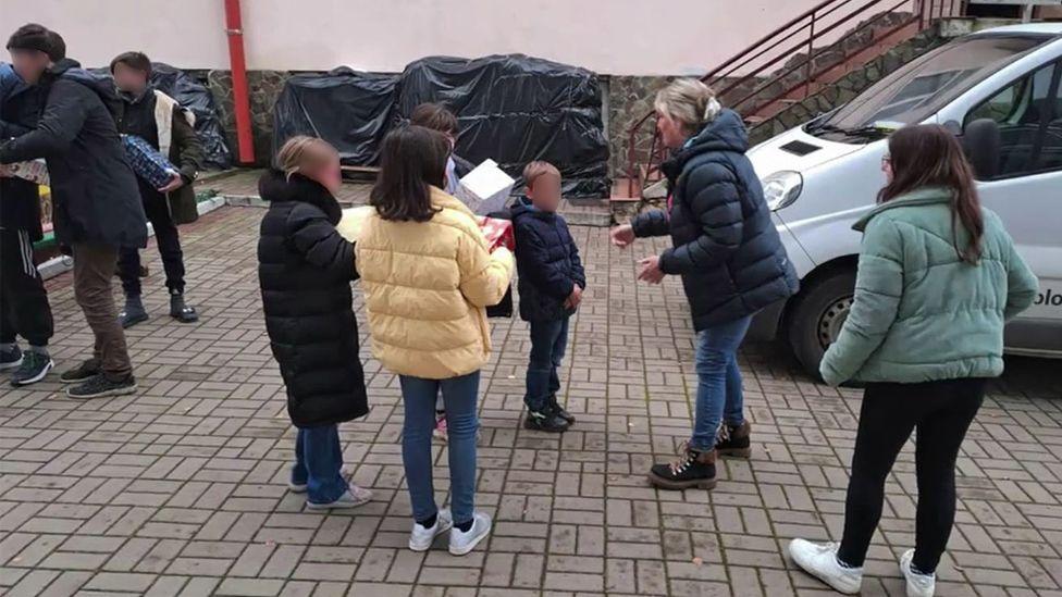 Jeanette Rice stands to greet four children in Ukraine. All are wearing winter jackets and three of the children are holding Christmas boxes. 