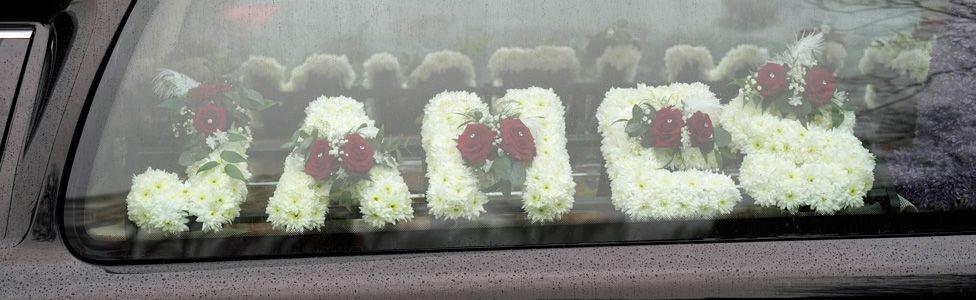 A floral tribute spelling "James" seen through a hearse window