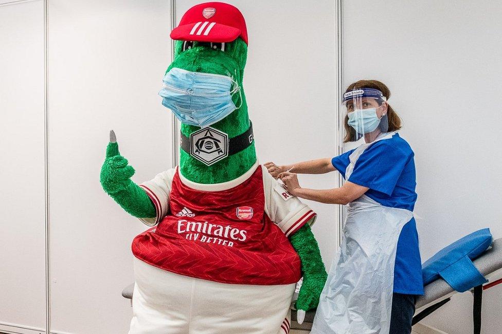 Arsenal mascot Gunnersaurus (left) gets his Covid vaccination from an NHS frontline worker