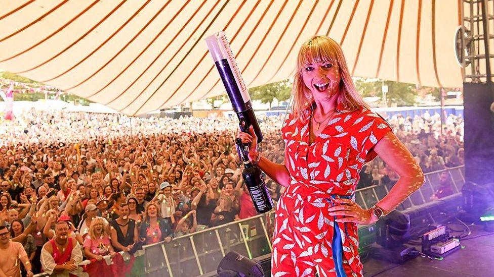 Sara Cox standing with her back to a festival crowd, grinning and holding a confetti cannon