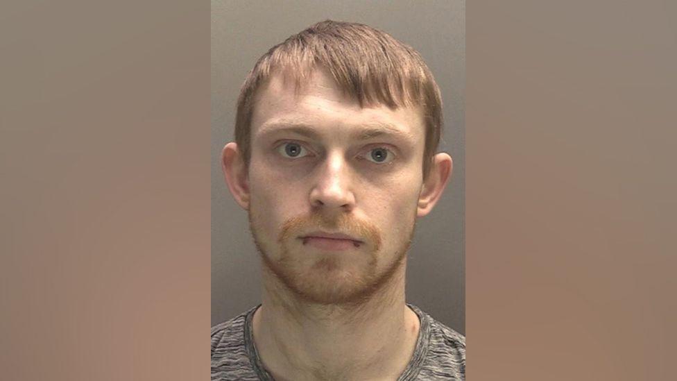 A police-issued mugshot of Kieran Williams. He has a neutral expression, fair short hair and a double bottom lip piercing. He is wearing a grey t-shirt.