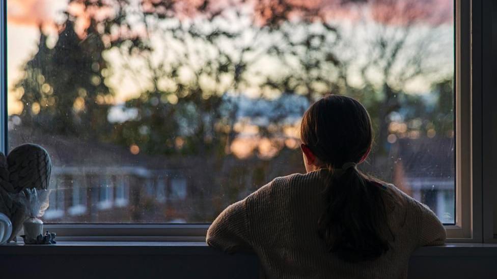 A young girl stares out of a window at dusk, with her elbows on the window ledge. There are properties in front with a backdrop of tall trees.