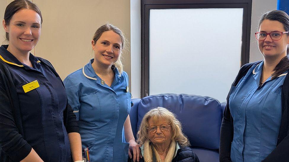 Nurses Danni Parkin and Jess Bratton in blue uniforms stand next to Anne who has grey hair and glasses and is sitting in a blue chair