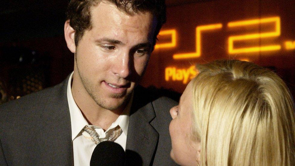 A young Ryan Reynolds, in a suit, looks down at an interviewer as she asks him questions. Both are standing in front of a sign showing the Playstation 2 logo in orange lettering