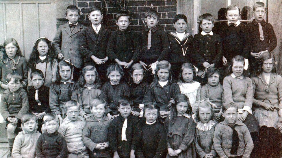 A black and white photo showing children at a primary school in about 1919. There are three rows of boys and girls. Behind them is a wall, door and window. 