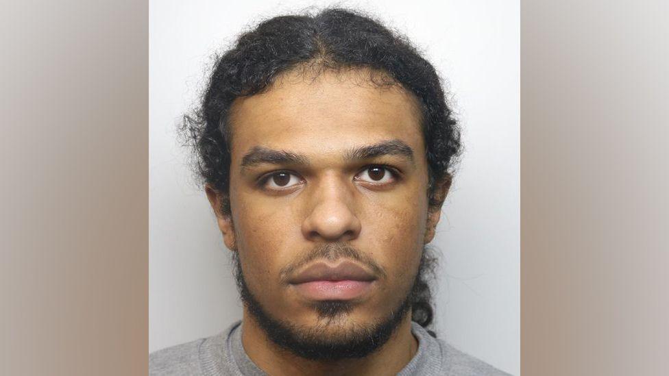 Jayden Gubetini mugshot pictured looking straight at the camera, not smiling. He has a goatee beard and hair tied back in a ponytail, brown eyes and wearing a grey sweater.