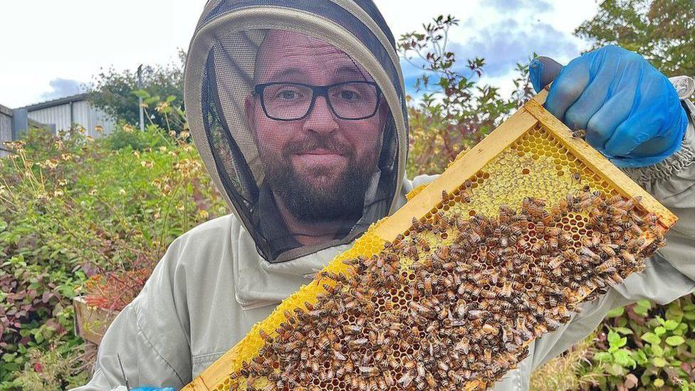 Ross Main standing in full anti-sting clothing holding up a honey hive full of bees.