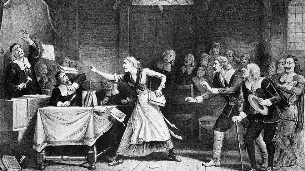 A black and white lithograph by George H Walker shows a witch trial in Salem, Massachusetts. A woman is standing in a room surrounded by men and books appear to be flying from her hands towards a judge