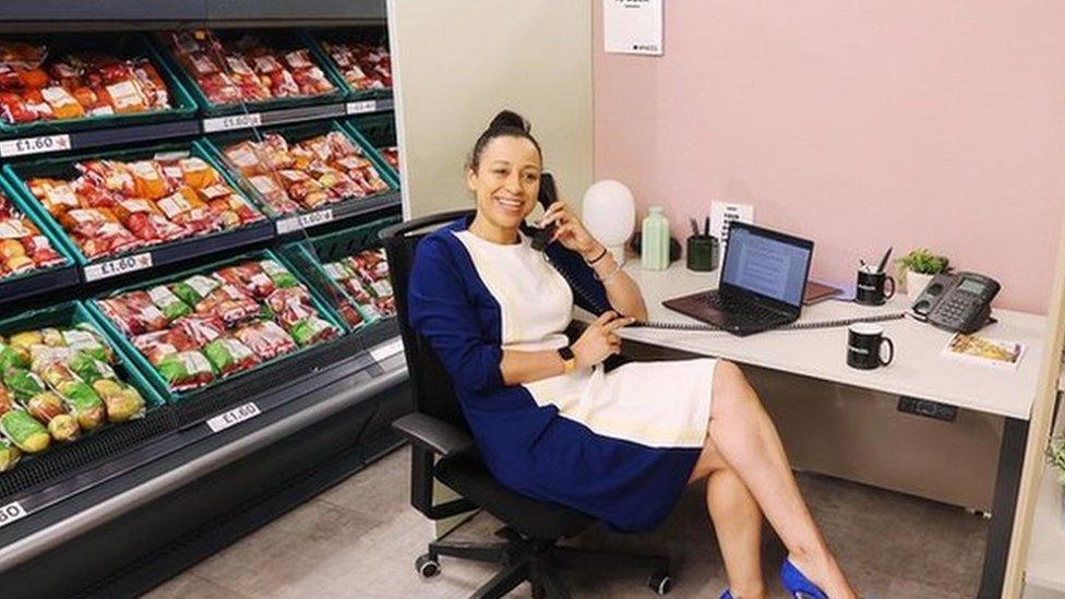 Woman sits at desk in the middle of a fruit and veg supermarket aisle